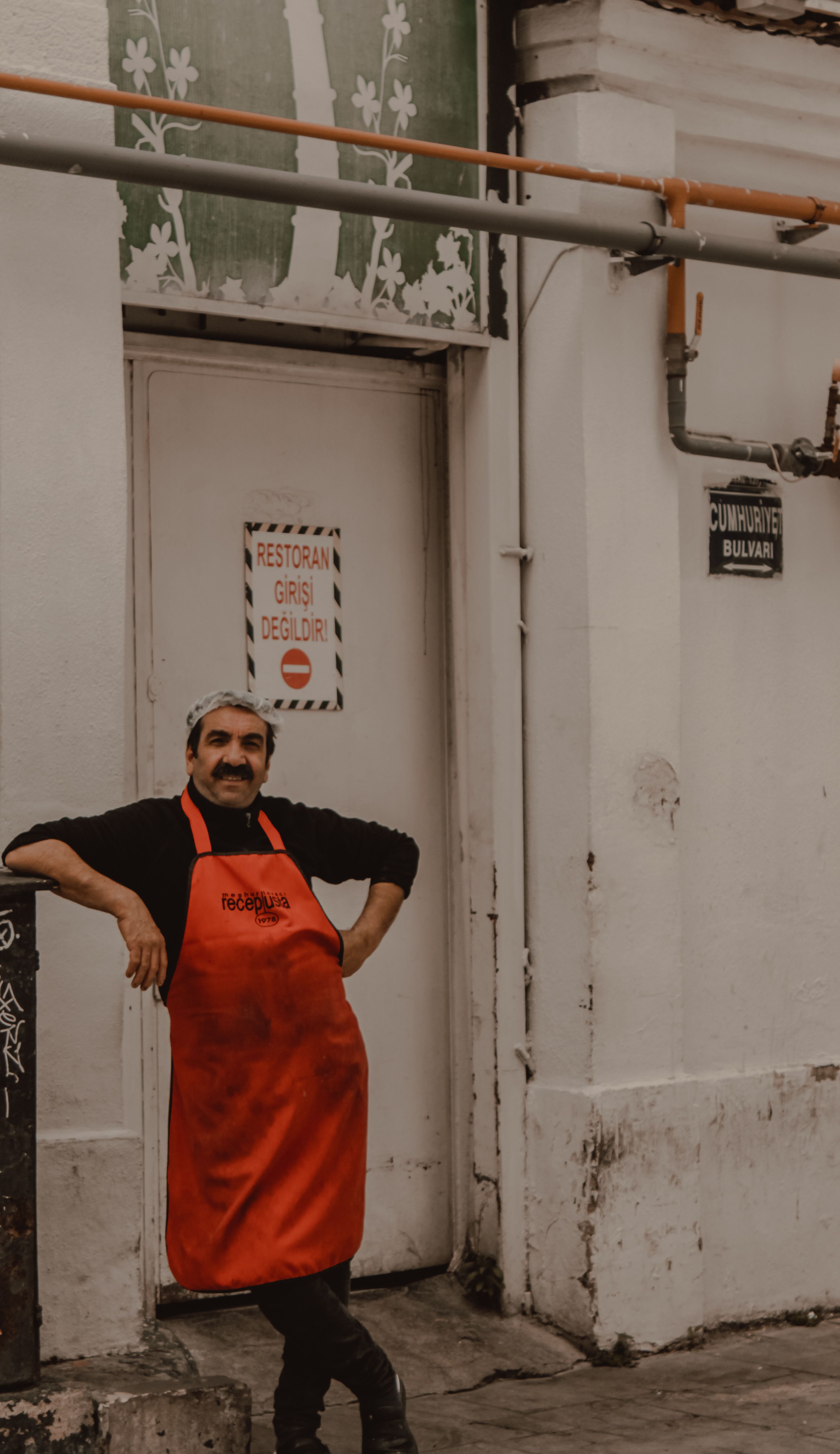 Candid shot of a chef in red apron leaning against a wall in a city alley.