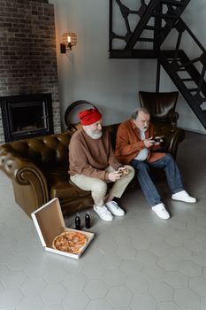 Two senior men playing video games on a couch with pizza and drinks nearby.