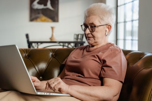 Free Senior adult woman with glasses using a laptop on a comfortable couch indoors. Stock Photo