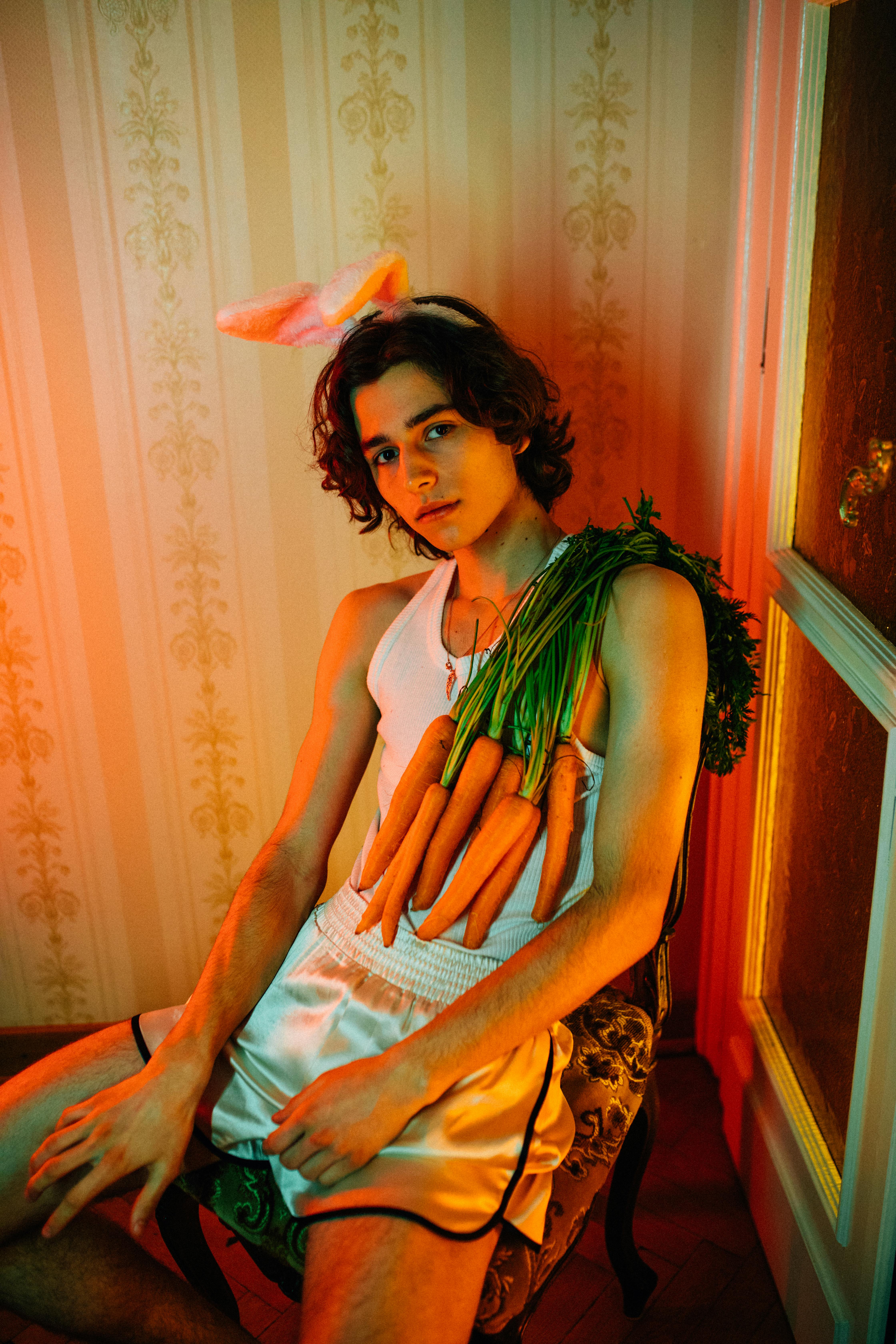 Teen model posing indoors with carrots and bunny ears, showcasing trendy fashion.