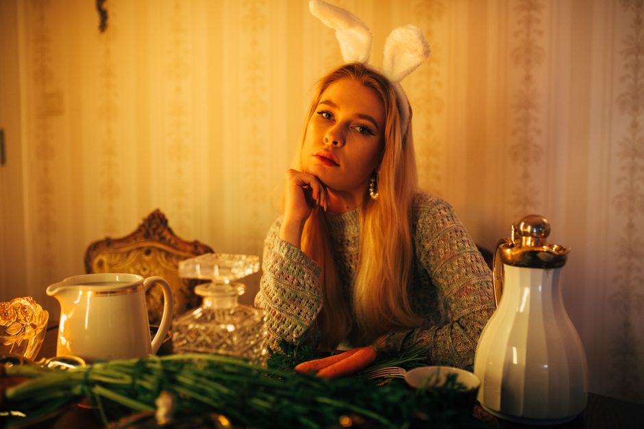 A woman with bunny ears poses thoughtfully indoors, surrounded by vintage decor.