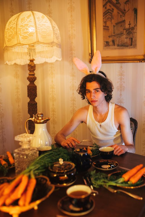 Free A Man Wearing Bunny Headband Sitting on a Chair Near the Wooden Table with Foods while Seriously Looking at the Camera Stock Photo