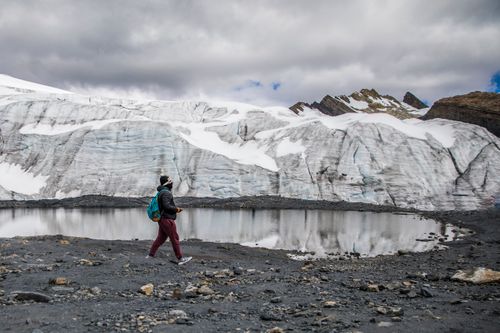 Free A lone hiker walks beside a glacier and a reflective pond under cloudy skies. Stock Photo