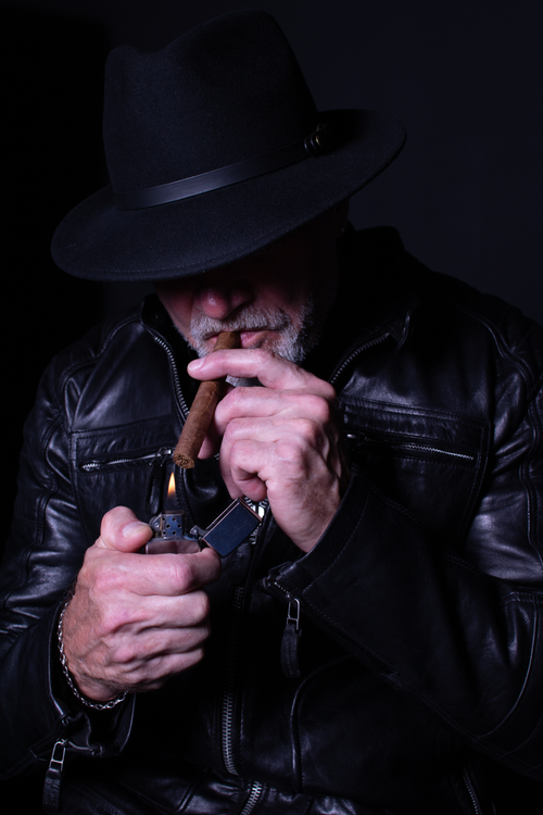 Free Senior man in leather jacket and hat lighting a cigar in a dark setting. Stock Photo