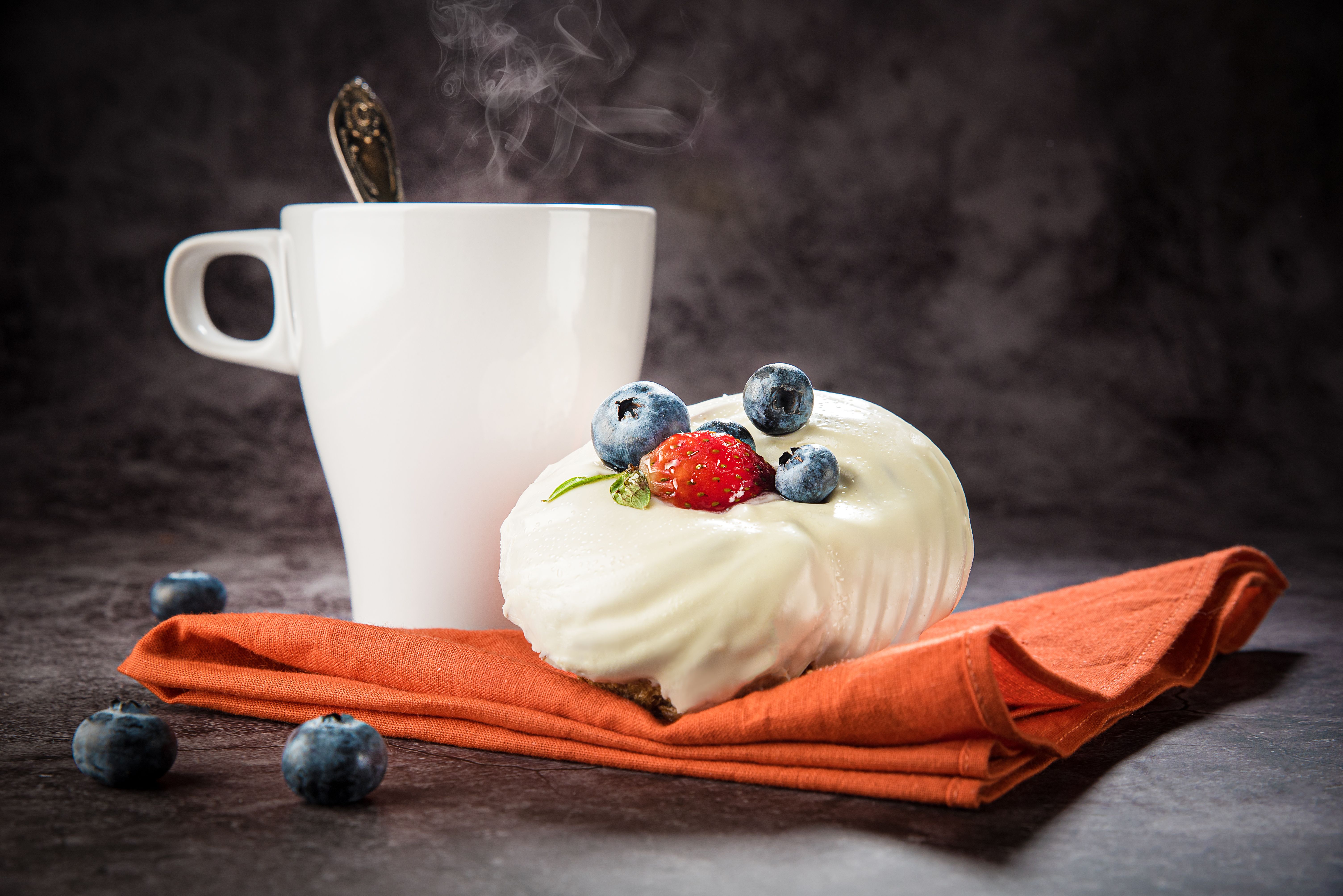 Imagem de uma xícara de café com uma colher e um doce com cobertura de creme, decorado com frutas vermelhas, sobre uma toalha laranja, sugerindo um momento de pausa e apreciação na rotina matinal.