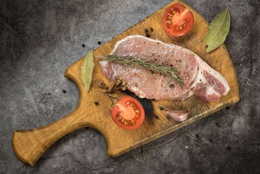Top view of a marinated raw pork steak with herbs and tomatoes on a wooden chopping board.