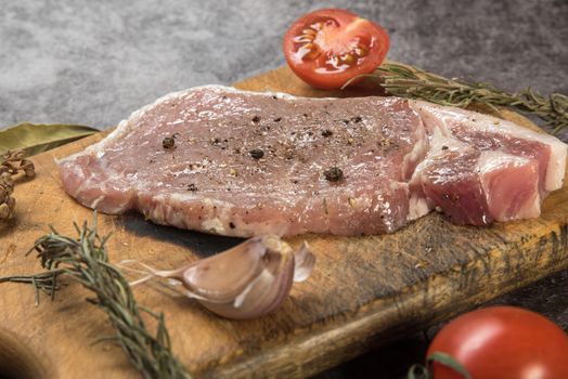 Fresh raw pork chop seasoned with peppercorns on wooden board surrounded by garlic, tomatoes, and herbs.
