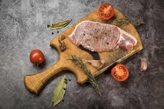 A raw pork steak with spices, herbs, and tomatoes on a wooden board for food preparation.