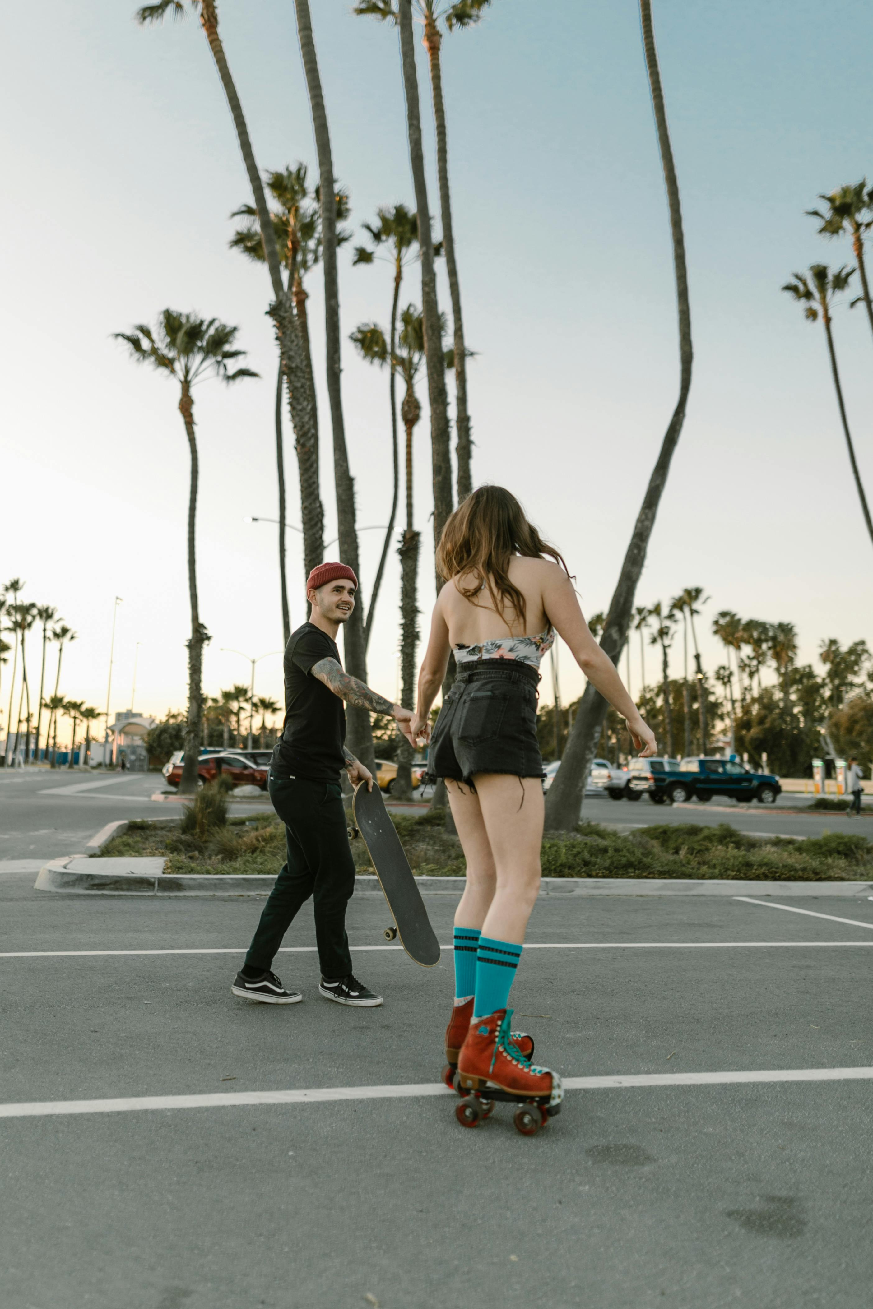 A young couple enjoys roller skating and skateboarding in a palm tree-lined urban park during the day.