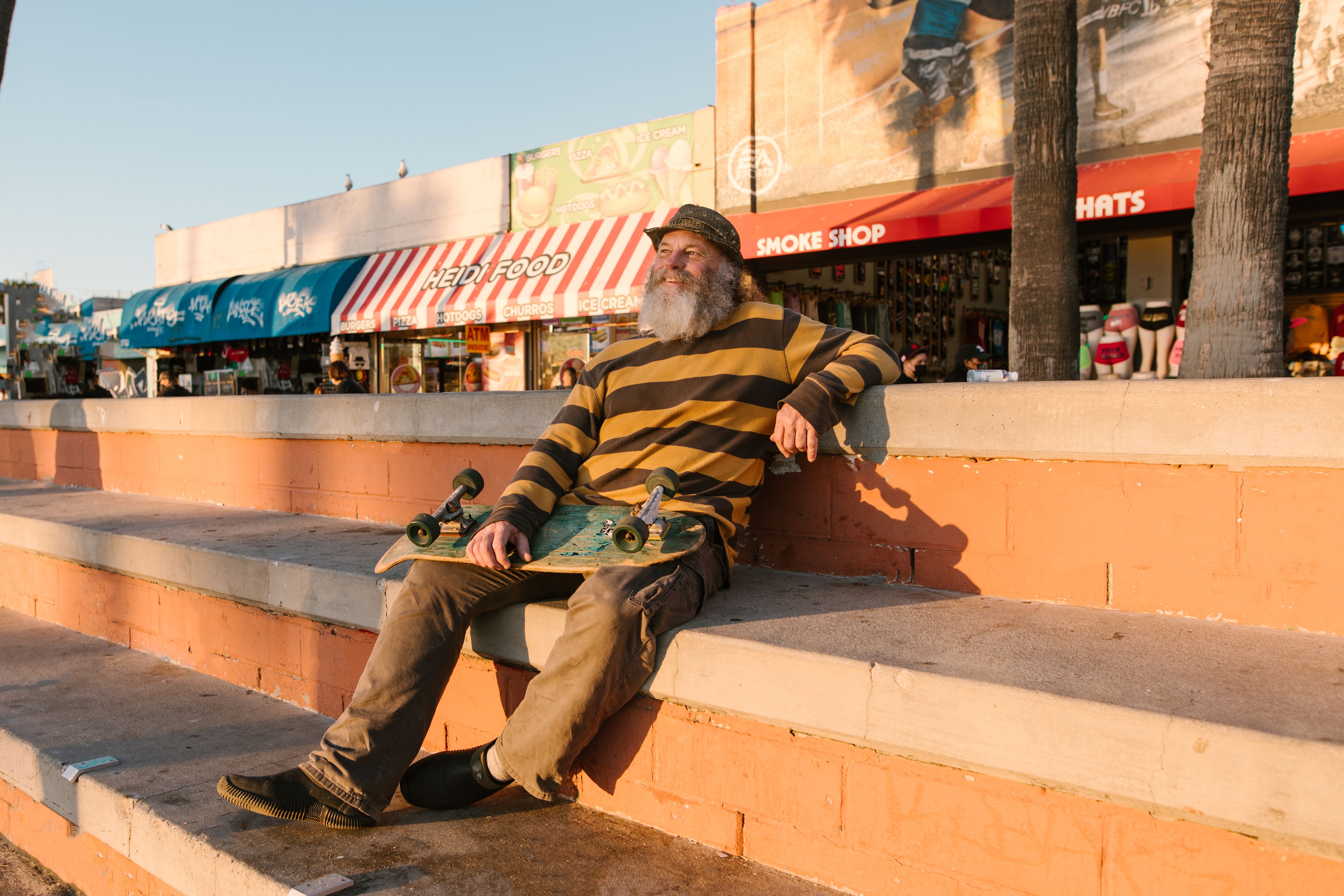 Bearded man sitting on steps with skateboard, enjoying sunny day outdoors.