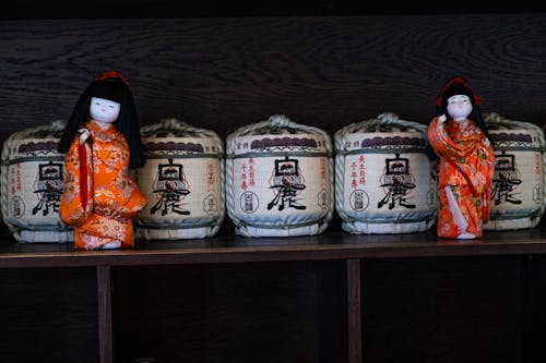 Free Traditional Japanese Kimekomi dolls in red kimonos placed on shelf with abundance of wooden sake barrels with ropes and hieroglyphs Stock Photo