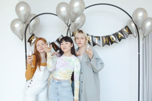 Three women celebrating a birthday with champagne indoors amidst balloons and decorations.