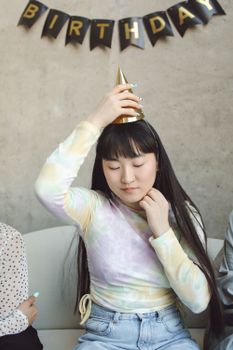 A young woman wearing a colorful top adjusts her party hat during a birthday celebration indoors.