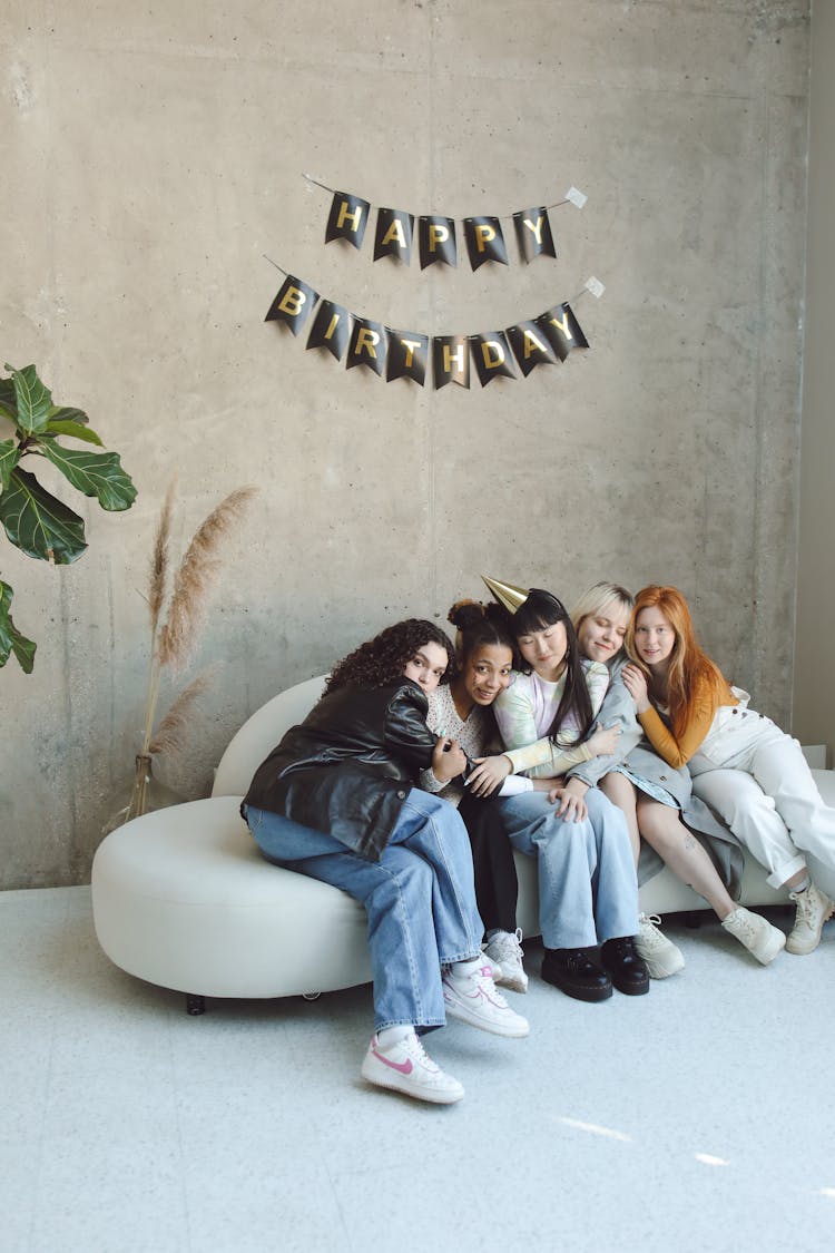 Group Portrait Of Young Women At A Birthday Party Sitting On A White Sofa Against Concrete Wall