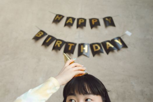 A young girl adjusts her party hat under a 'Happy Birthday' banner indoors.