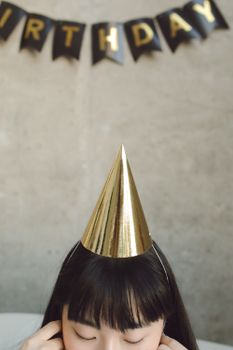 Asian woman wearing a golden party hat at a birthday celebration, eyes closed.