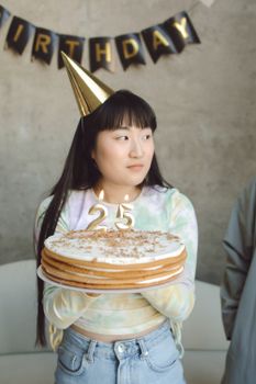 Asian woman holding a cake with 25 candles at her indoor birthday celebration.