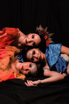 Three women adorned with glitter makeup and colorful costumes pose elegantly in a studio setup.