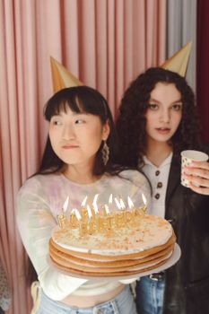 Two women celebrate a birthday with a cake and party hats, enjoying a fun indoor party.