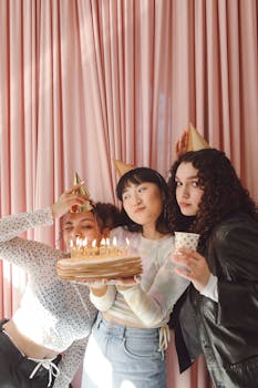 Three friends celebrating a birthday with cake, candles, and party hats against a pink backdrop.