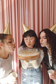 Three friends celebrate a birthday indoors, wearing party hats and blowing out candles on a cake.