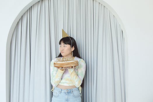 A young woman holding a birthday cake with decorations, wearing a party hat indoors.