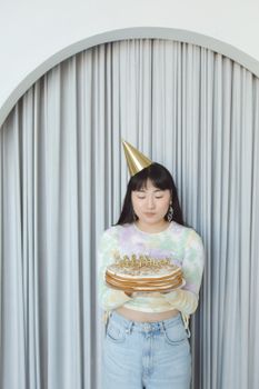 Asian woman in birthday hat holding celebratory cake indoors.