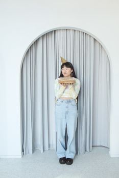 Young woman wearing a birthday hat holds a celebration cake in a minimalist indoor setting.