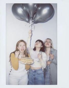 Three young women celebrate indoors with balloons and cake in a casual setting.