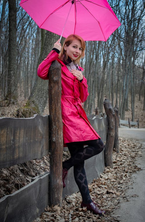 Young stylish female wearing trendy pink raincoat leaning on wooden fence while holding umbrella overhead while standing on walkway in autumn park