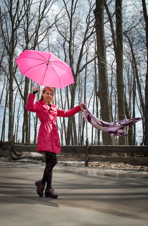 Free Happy smiling female standing on asphalt road in autumn park while holding umbrella over head and scarf flying in wind and wearing pink outerwear Stock Photo