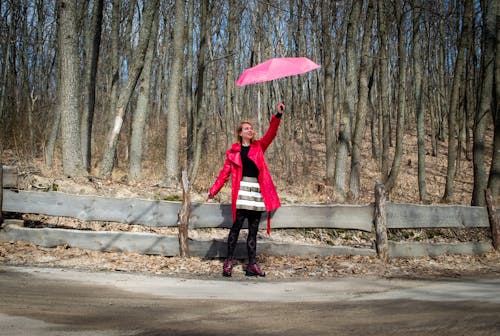 Optimistic young woman raising pink umbrella smiling while standing near wooden fence on road with leafless trees on background on sunny autumn day