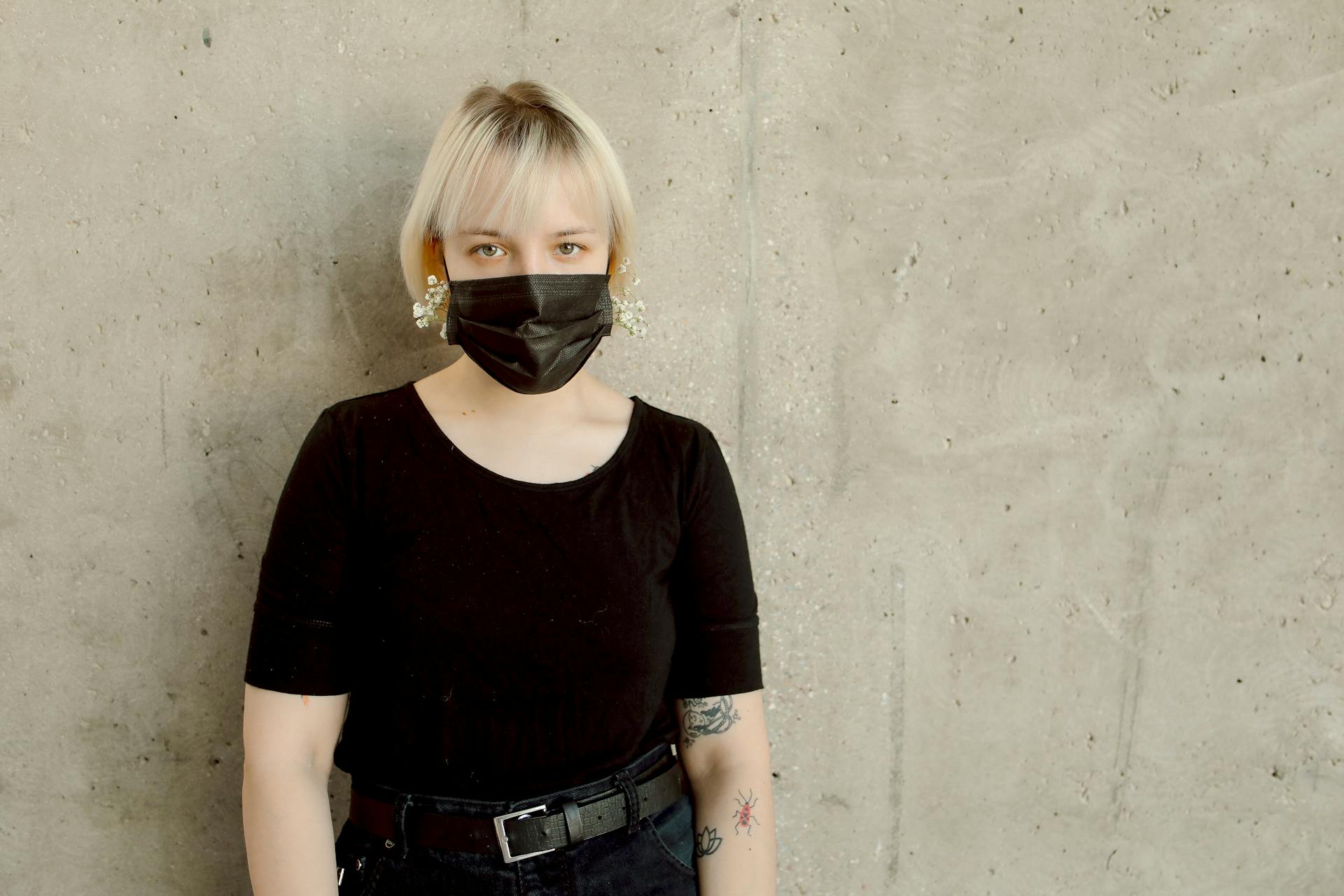 Blond woman wearing a black mask and black shirt against a concrete wall.