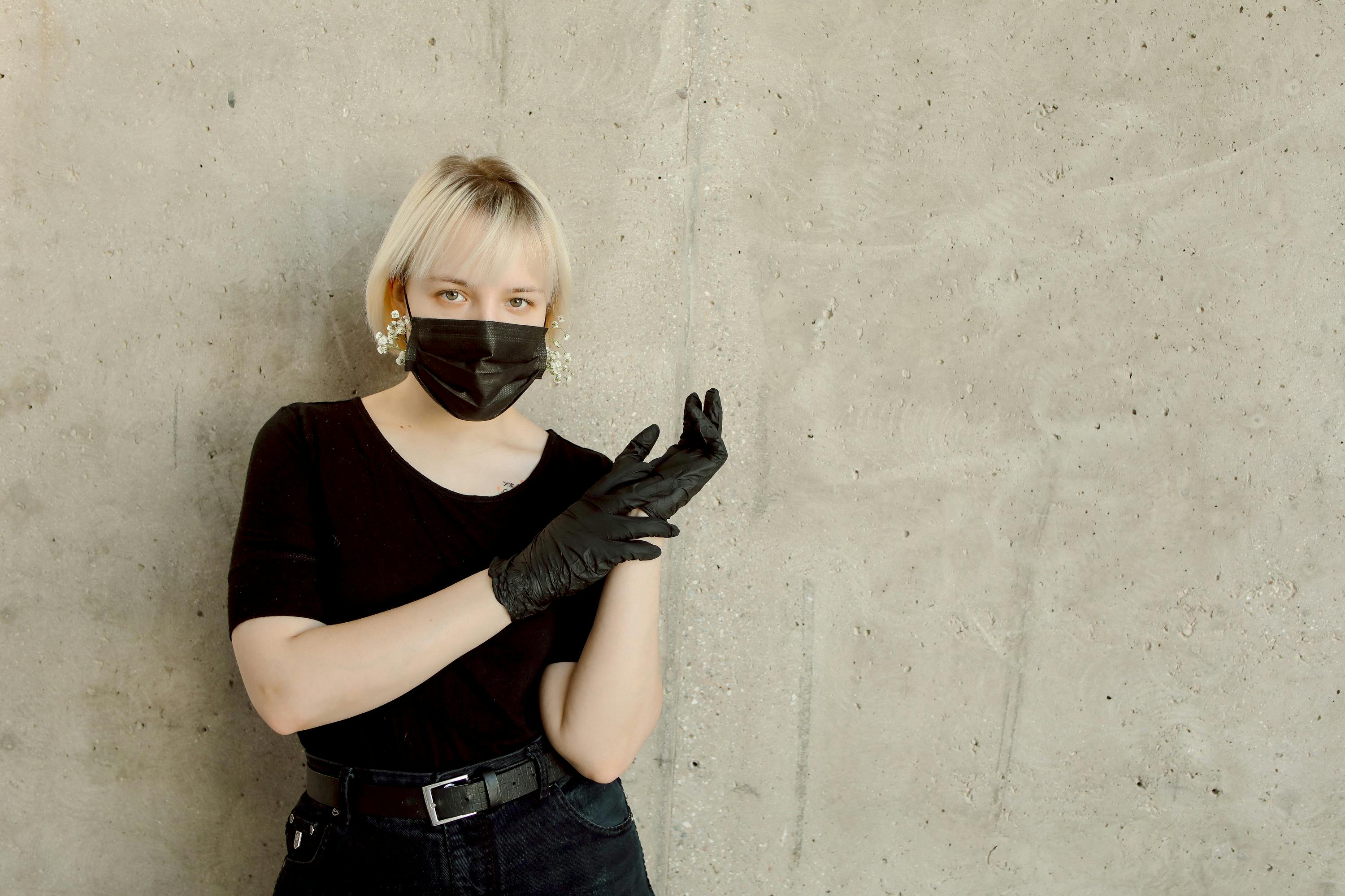 Caucasian woman with blond hair wearing a black mask and gloves against a concrete wall.