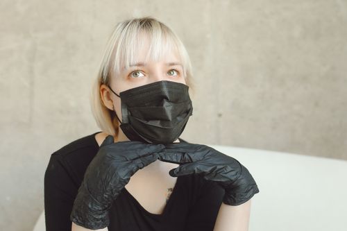 Free Caucasian woman with blonde hair wearing a black mask and gloves, gazing upwards thoughtfully indoors. Stock Photo