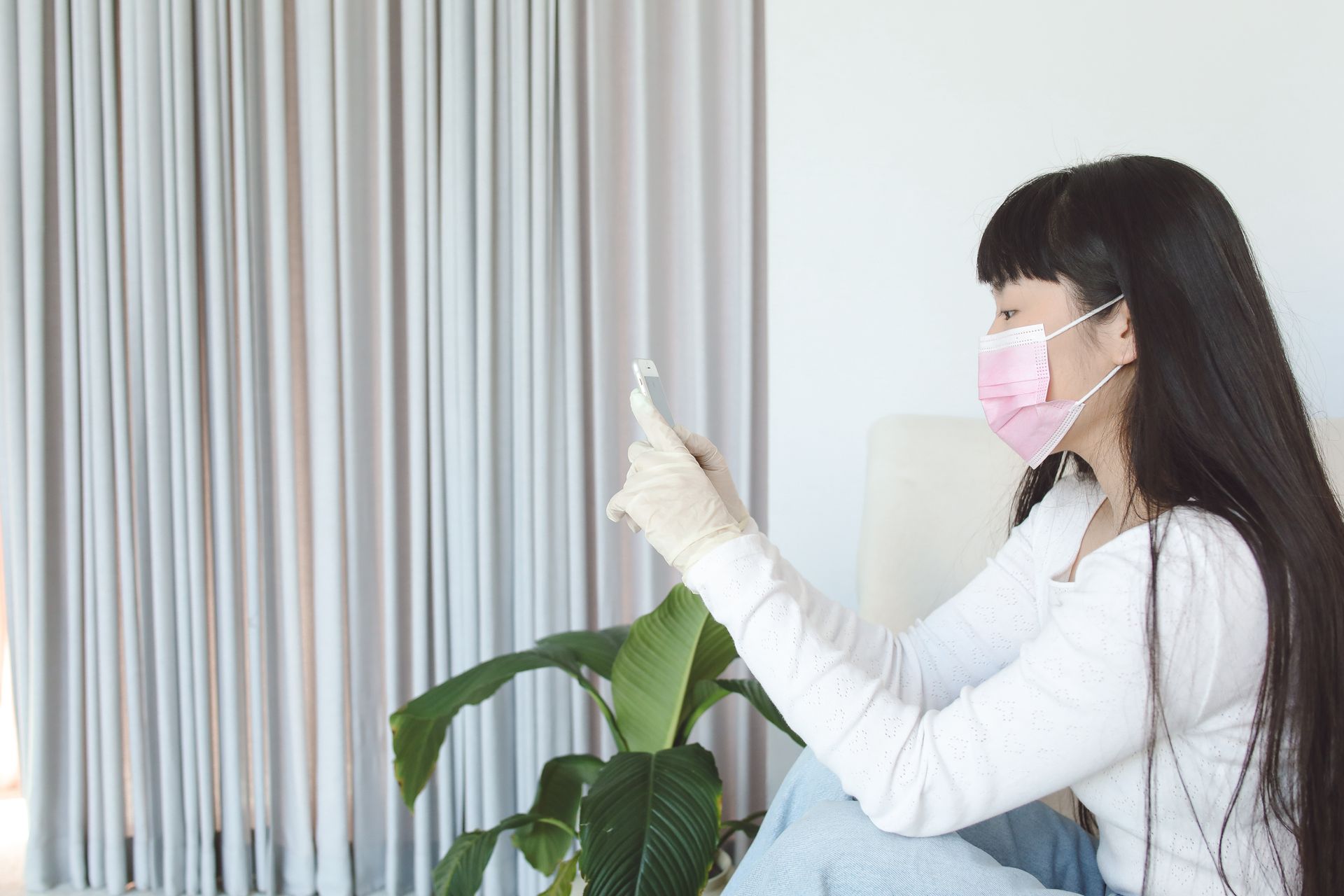 Asian woman in mask with smartphone indoors, practicing pandemic safety.