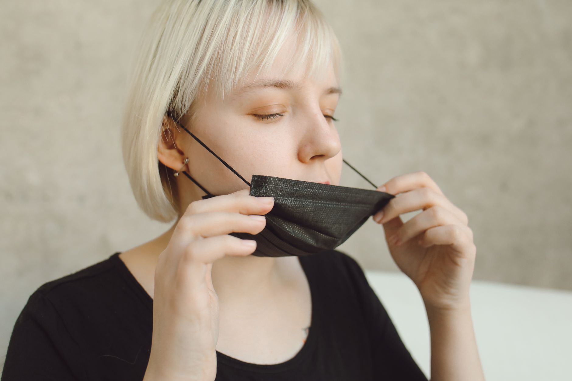A woman with short blonde hair adjusts her black face mask indoors for safety.