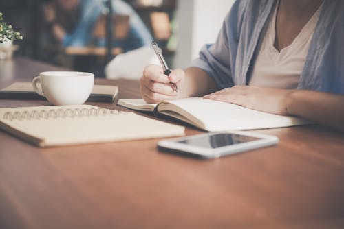 Free Woman Writing on a Notebook Beside Teacup and Tablet Computer Stock Photo