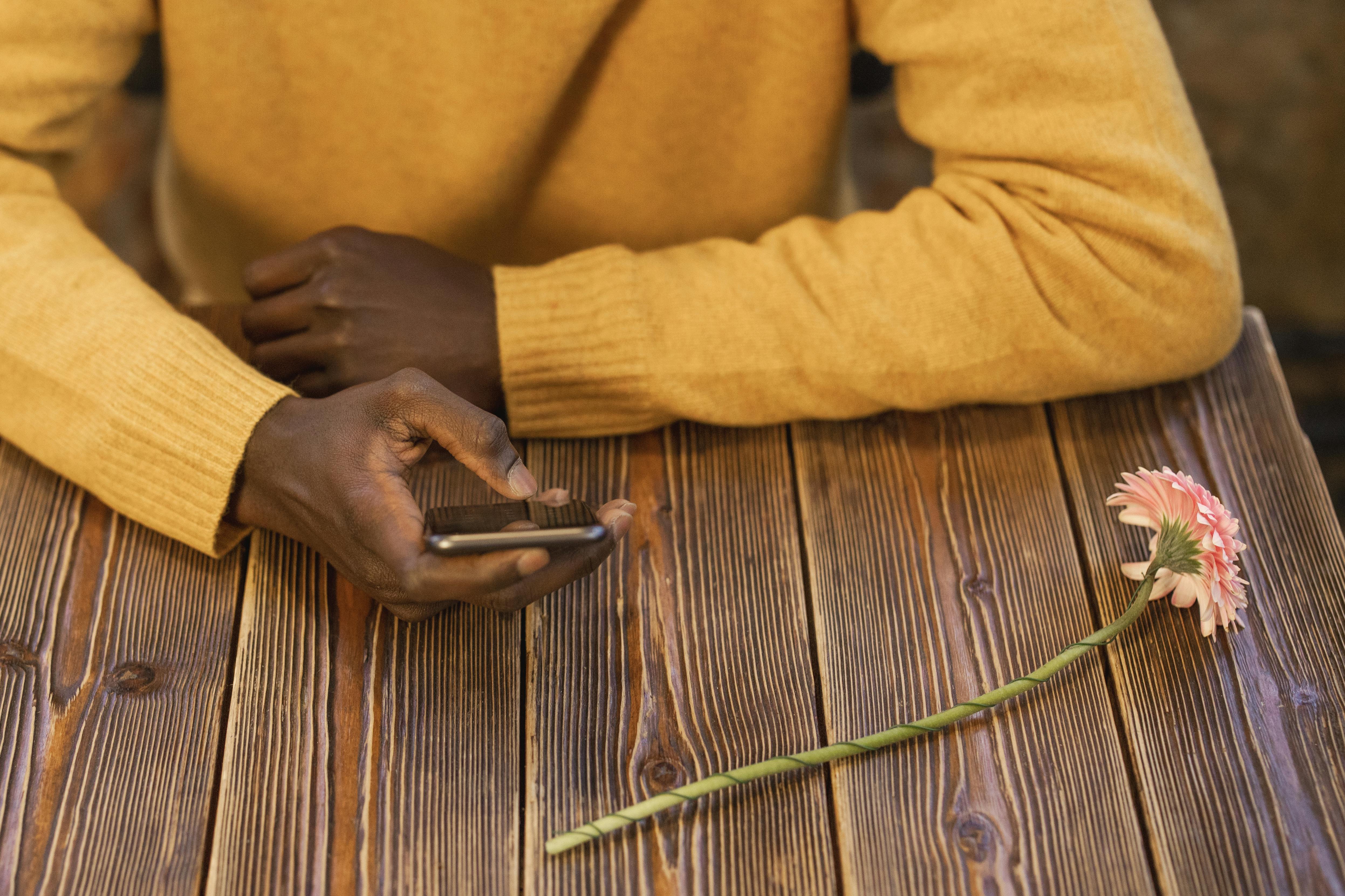 A person in a yellow sweater holds a smartphone on a rustic wooden table beside a single flower.