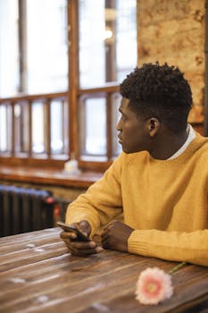 African American man in yellow sweater using smartphone and looking away at a cozy café table.