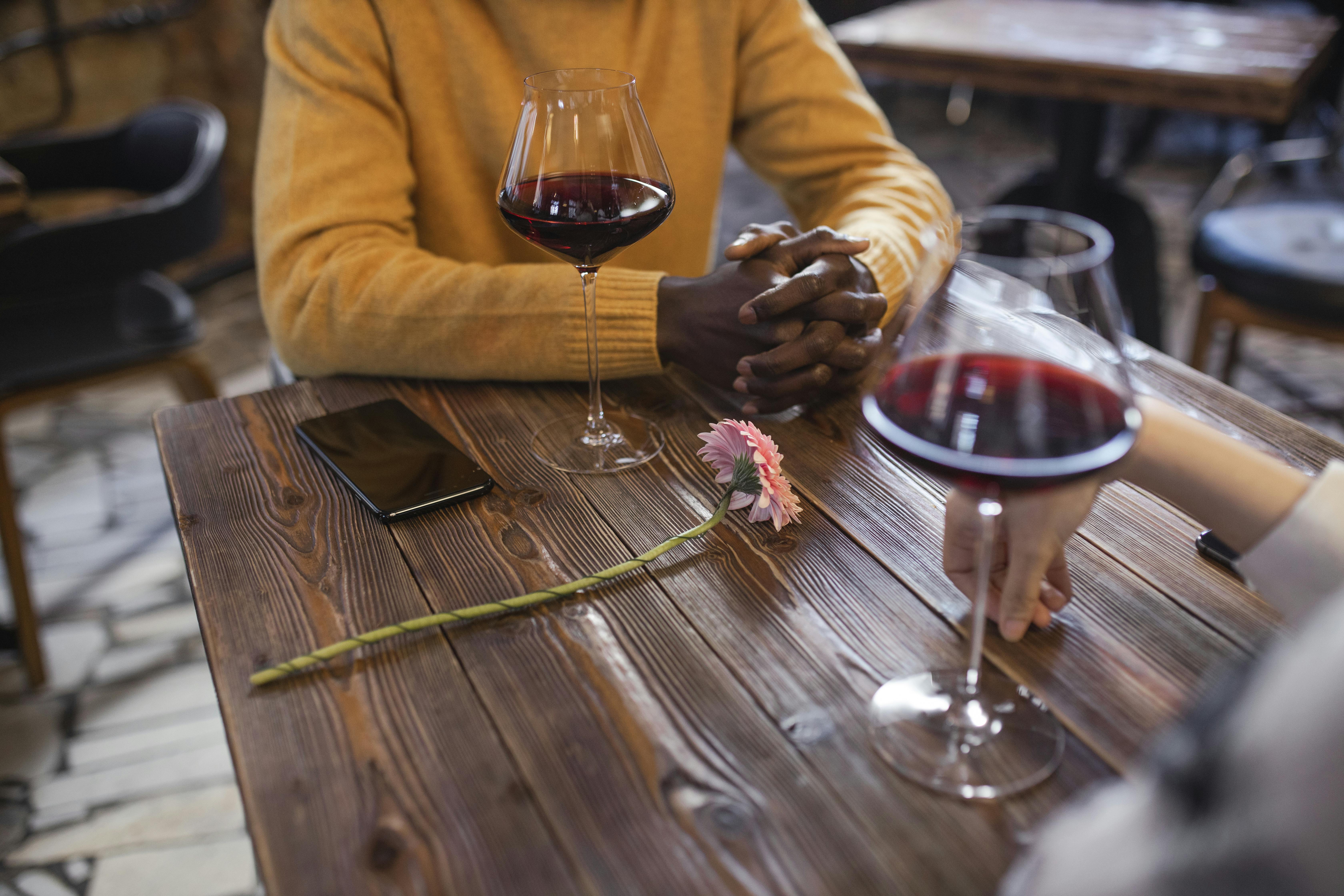 People enjoying red wine and a flower on a rustic wooden table in a cozy cafe setting.