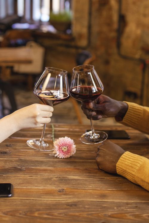 Free Two people toast with red wine glasses over a rustic table with a pink flower. Stock Photo