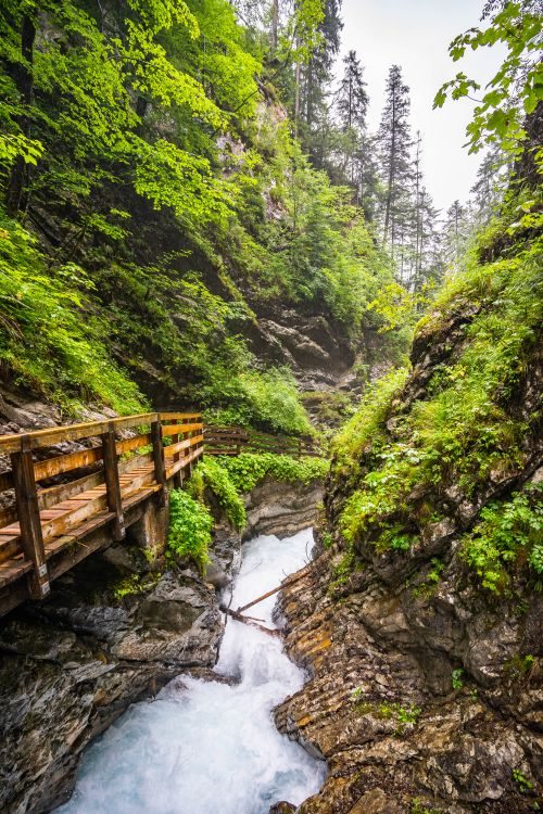 Free A picturesque view of a wooden bridge crossing a flowing stream amidst lush greenery in a mountainous area. Stock Photo