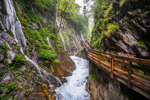Free A picturesque view of a wooden bridge crossing a vibrant stream with cascading waterfalls in a lush forest. Stock Photo