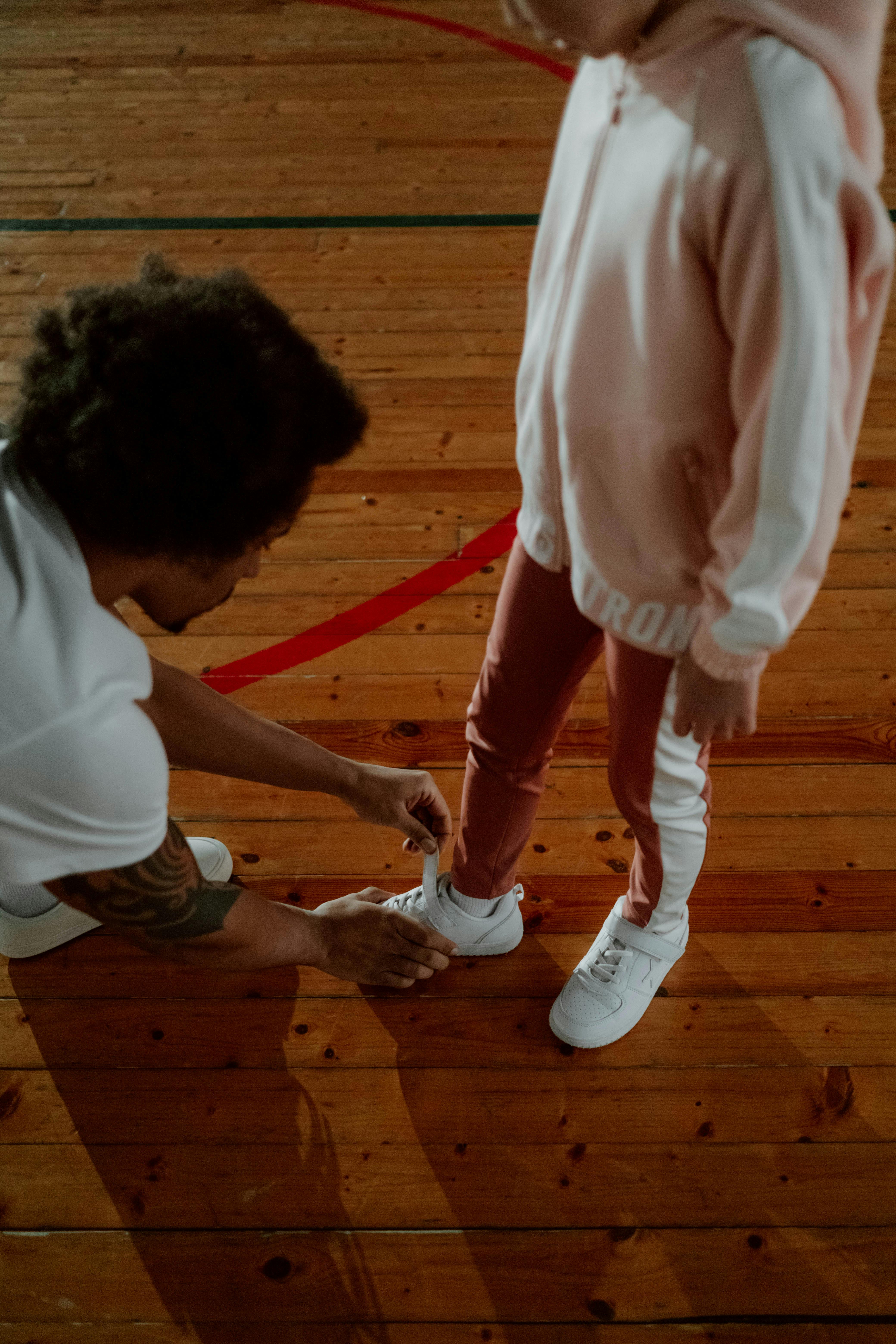 A man helps a child tie their shoes on a wooden gymnasium floor, fostering assistance and care.