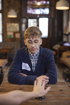 Young man with eyeglasses sitting indoors during a speed dating event, wearing a plaid shirt and blazer.