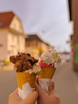 Two ice cream cones with various flavors held outdoors in a sunny street scene.