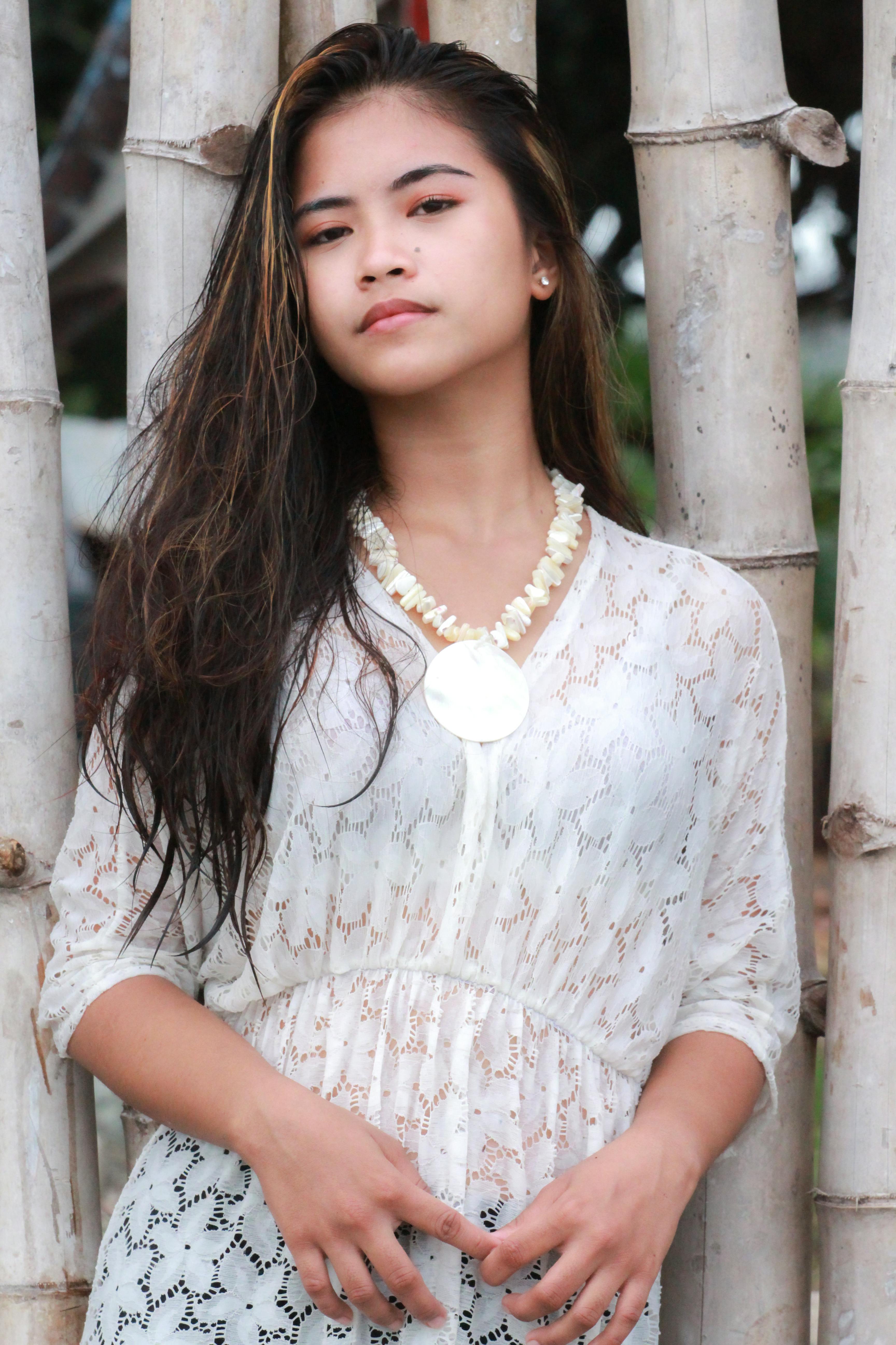 Portrait of a young woman in a white lace dress with a necklace, standing against bamboo.
