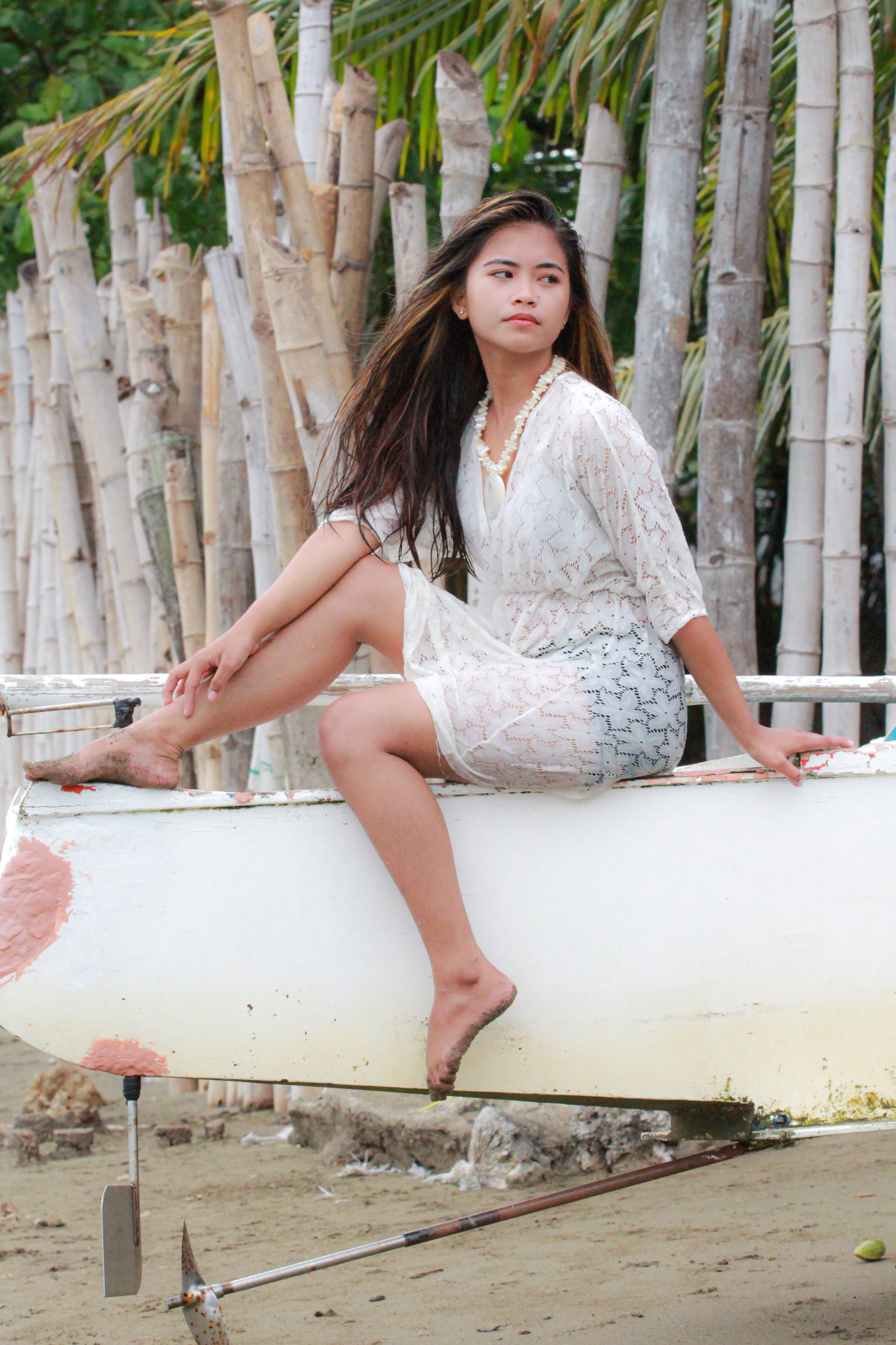 A young woman in a sheer dress poses on a canoe by the beach, surrounded by bamboo.