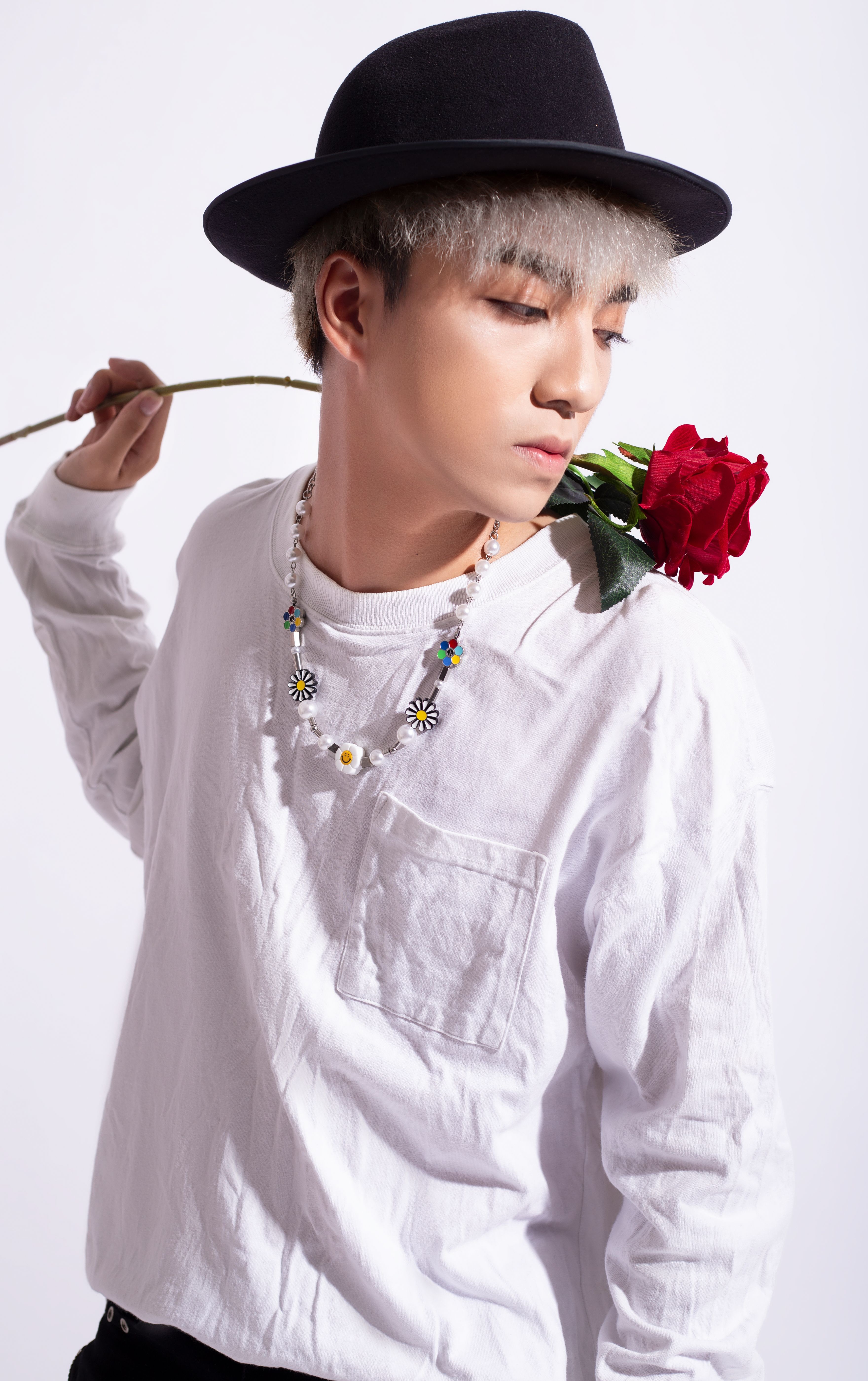 A stylish man in a hat poses with a rose on his shoulder in a studio portrait.